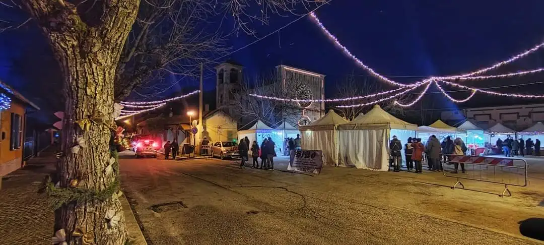 Tendoni del mercatino e luminarie sospese davanti a una chiesa in pietra con campanile, di notte - mercatini di Natale di Alba Fucens