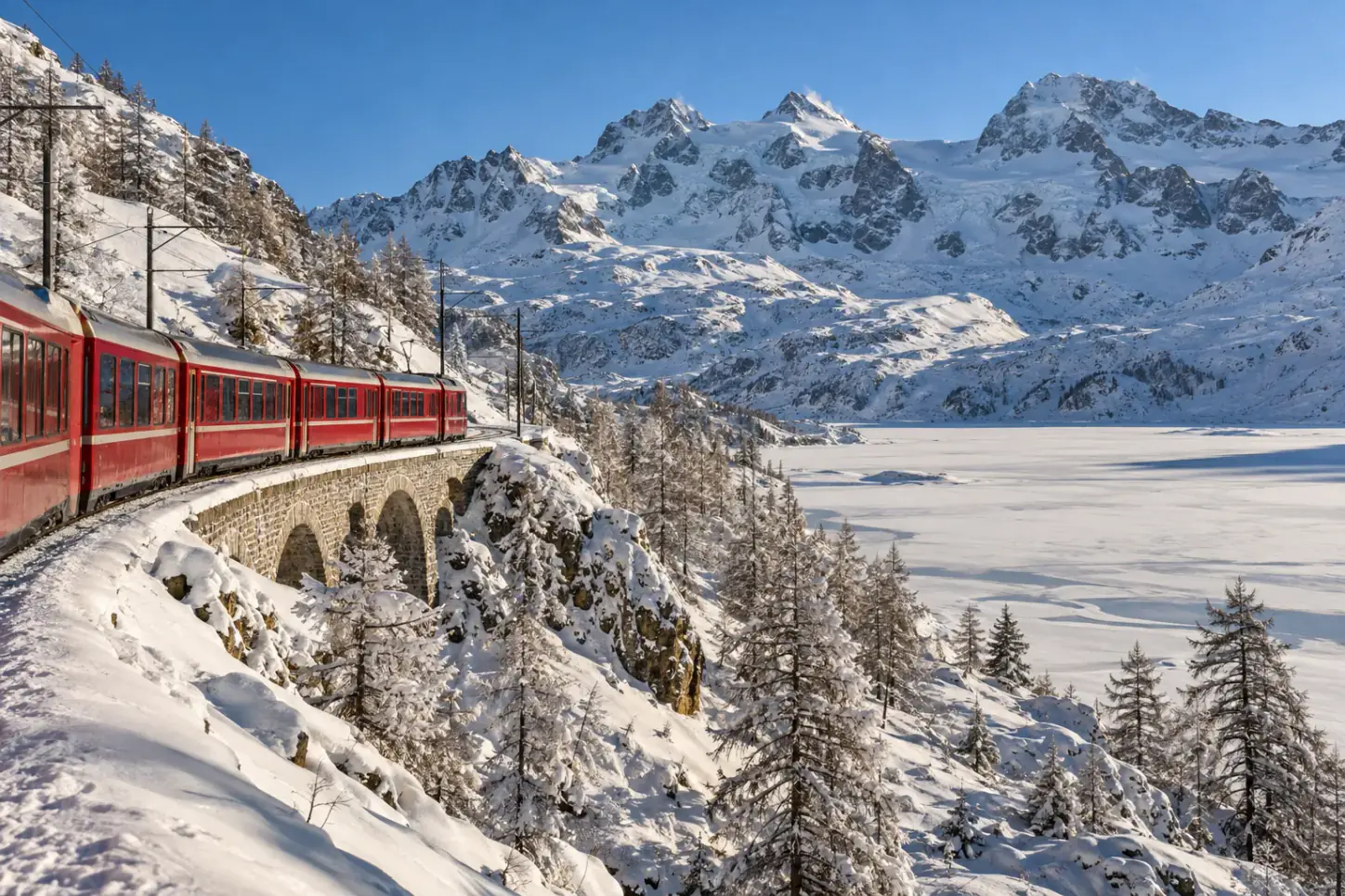 Il Trenino Rosso del Bernina su un viadotto in pietra tra boschi e vette innevate, in una giornata invernale limpida